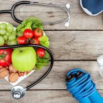Healthy lifestyle concept with fruits, vegetables, nuts in heart-shaped bowl, stethoscope, blue sneakers, jump rope, and water bottle on wooden surface.