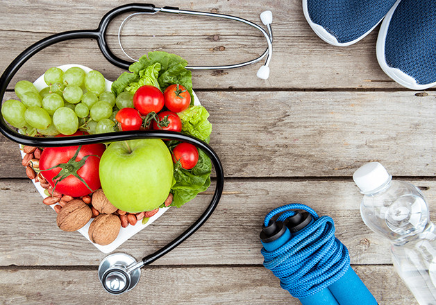 Healthy lifestyle concept with fruits, vegetables, nuts in heart-shaped bowl, stethoscope, blue sneakers, jump rope, and water bottle on wooden surface.