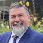 Smiling man with a beard wearing a blue suit and patterned tie, standing outdoors with greenery in the background.