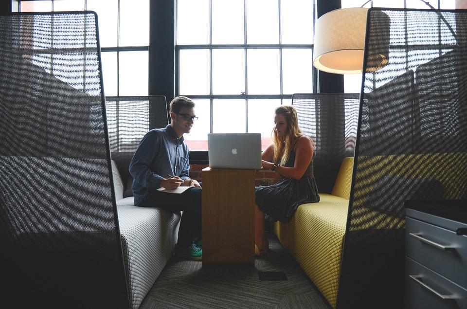 Two people sit in a modern office booth, working together on a laptop. A lamp hangs overhead, and large windows are in the background.