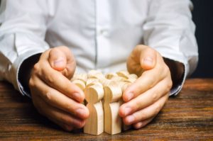 A person in a white shirt uses both hands to encircle several small wooden human figures on a wooden table.