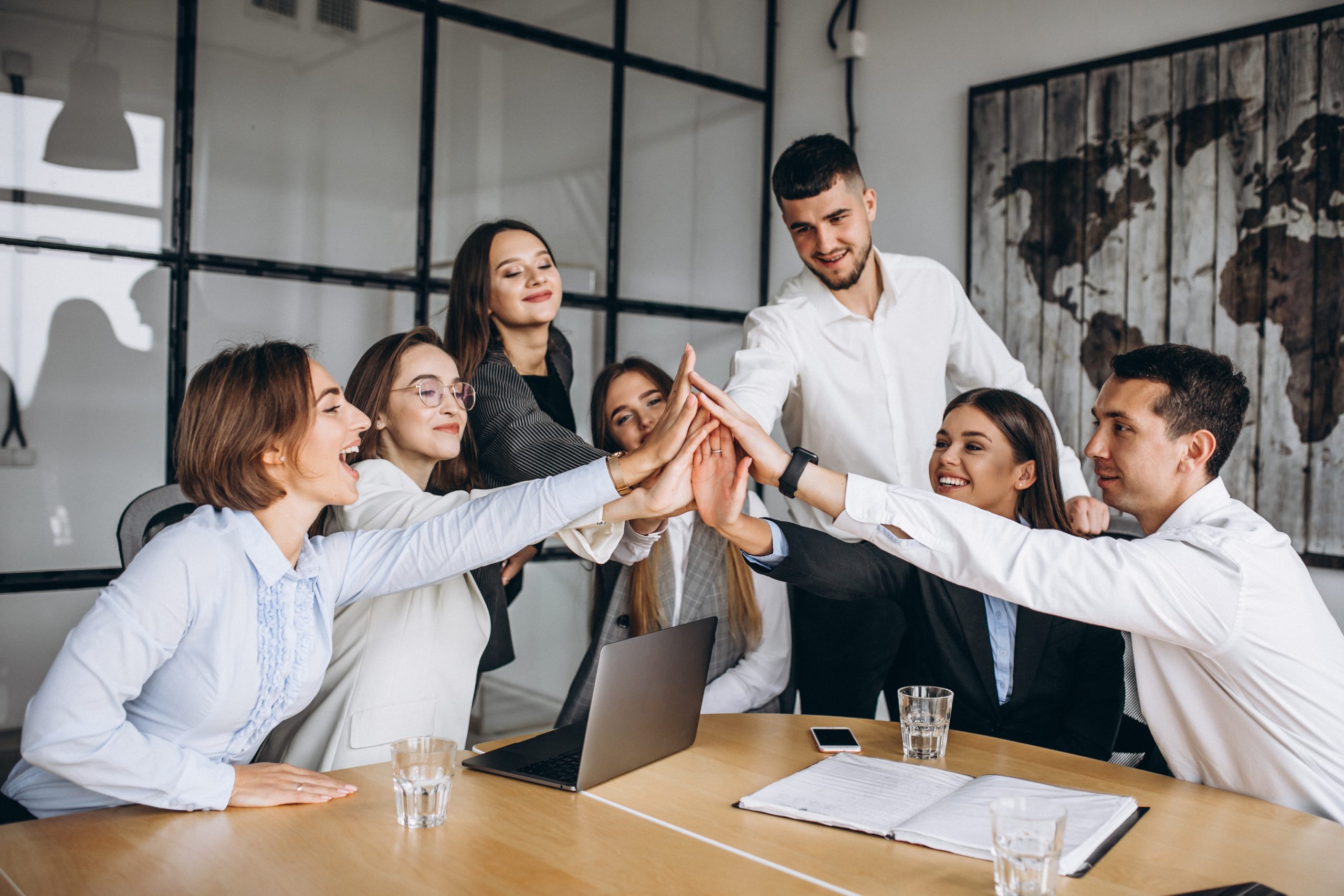 A group of seven colleagues in business attire gather around a table, reaching in for a collective high-five, with glasses of water and a laptop on the table.