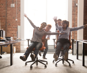 Office setting with two men pushing two women on office chairs having fun