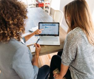 Two people sit at a table with a laptop, focused on enhancing their financial wellbeing. One person points at the screen displaying a webpage filled with insights. Papers and a notebook are scattered on the table, supporting their exploration of financial strategies.