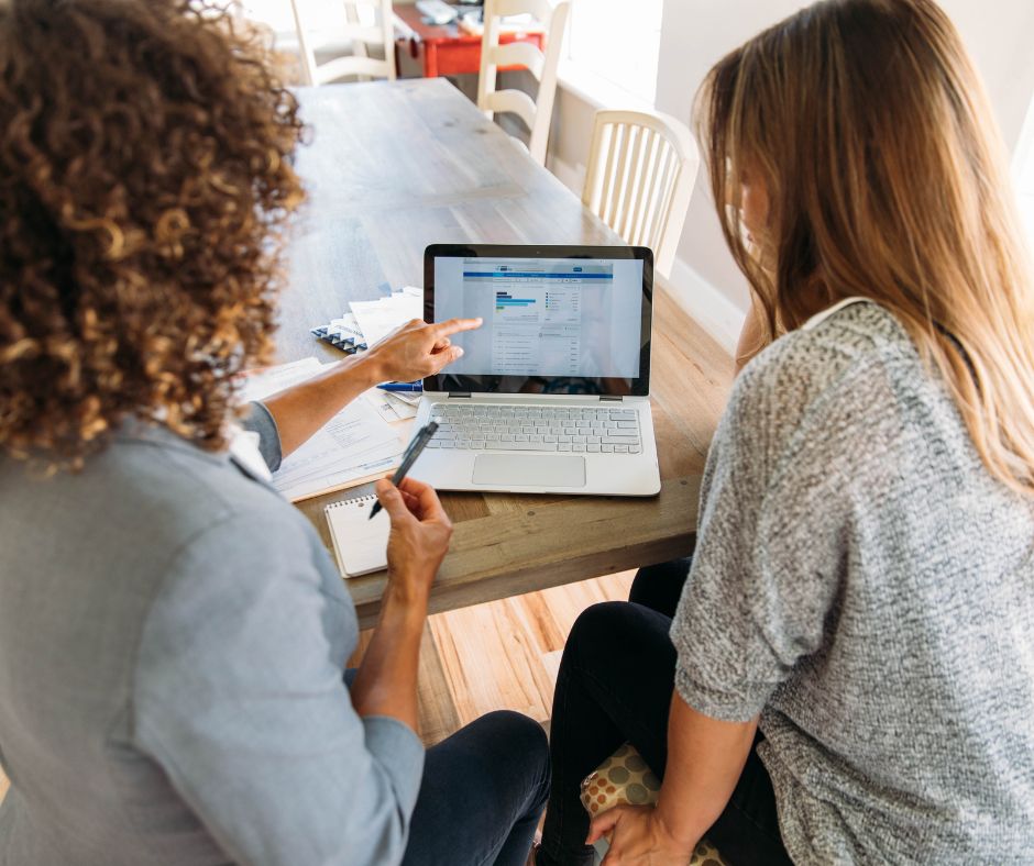 Two people sit at a table with a laptop, focused on enhancing their financial wellbeing. One person points at the screen displaying a webpage filled with insights. Papers and a notebook are scattered on the table, supporting their exploration of financial strategies.