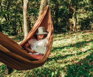 A person in a hammock, wearing a hat and holding an open book, enjoys the importance of annual leave while relaxing in a wooded area with dappled sunlight dancing on the grass.