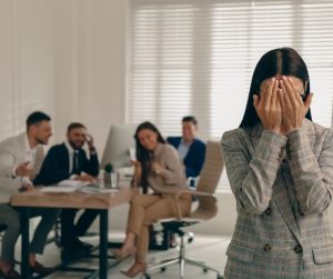 A woman covers her face in distress in the foreground, showcasing signs of a toxic work environment, while a group of four people in an office setting appear to be laughing or talking in the background.