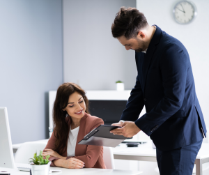 A man in a suit shows a tablet to a woman seated at a desk in a modern office, with a clock and plant visible in the background.