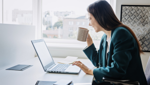 A woman in a blue blazer sits at a desk, smiling while looking at a laptop and holding a coffee mug, with windows showing a blurred cityscape in the background.