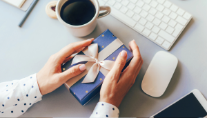Person holding a wrapped gift box with a ribbon, next to a coffee cup and computer keyboard on a desk.