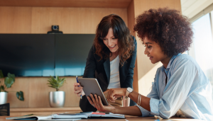 Two women collaborate at a table, one holding a tablet. There are books, plants, and a large screen in the background.