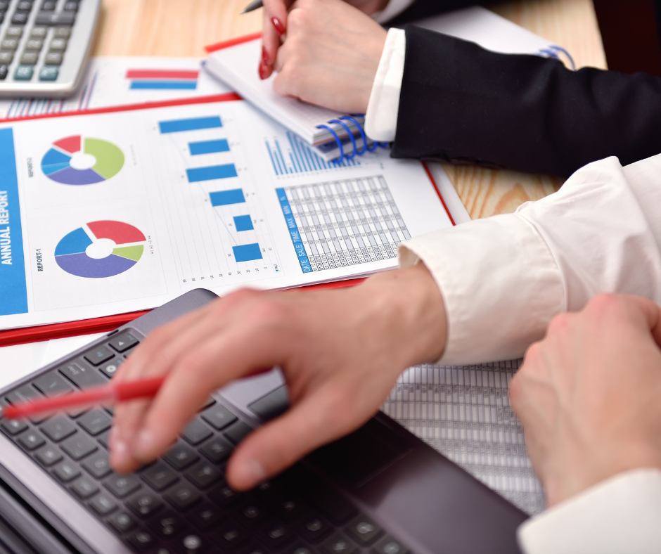 Two people in business attire review financial charts and graphs on printed reports while using a laptop and taking notes with a pen at a desk, discussing updates to the annual leave policy.