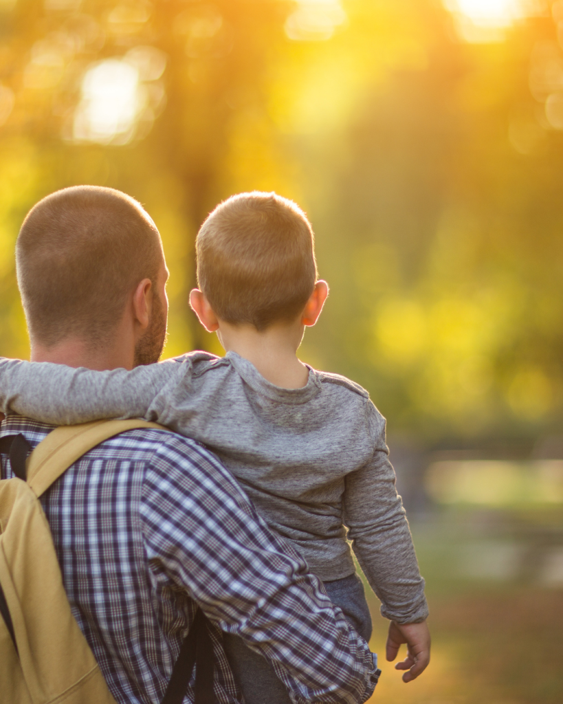 A father carries his young son through a sunlit park, capturing a peaceful moment made possible by flexible working hours and benefits for working parents.